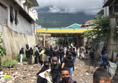 Masked students in a canal flanked by buildings, cleaning up garbage, with a yellow and blue bridge and the volcano Gamalama in the background