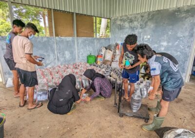 Three male students to the left pouring compost in a bag and weighing it, two female students to the left crouching and labeling the bags of compost, two male students further to the left handing the female students labels for the compost bags in a room filled with bags of compost
