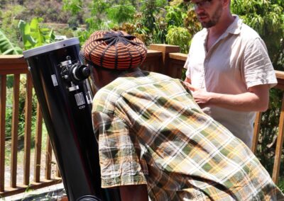Elderly bearded man in Jamaican garb peering through a telescope while Lev stands off to the side and talks