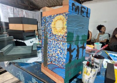 A rectangular wooden box pained with van Gogh's Starry Night on one side and the word "mel"and some cartoon trees on the other side sits on a table near paint