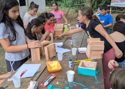 Multiple girls work on painting beehive boxes on a wooden table outside in the rain forest