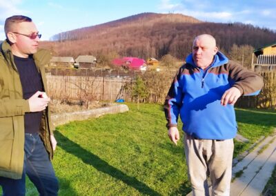 Two men stand in a field talking, with farms and mountains in the background