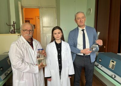 An older gentleman in a lab coat holding a closed jar with water, a young woman in a lab coat, and another older gentleman in a tie and sweater holding a flask with water